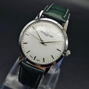 Pu&ograve; includere: Un orologio Jaeger-LeCoultre Club argentato con quadrante bianco e cinturino in pelle verde. L'orologio ha lancette e indici argentati, e le parole "SWISS MADE" sono stampate sul quadrante. L'orologio &egrave; esposto su un supporto trasparente.