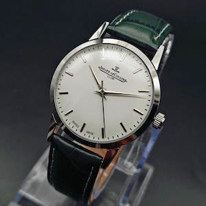 Pu&ograve; includere: Un orologio Jaeger-LeCoultre Club argentato con quadrante bianco e cinturino in pelle nera. L'orologio ha un quadrante rotondo con lancette e indici argentati. Il cinturino &egrave; nero con cuciture bianche e un cinturino in pelle verde. L'orologio &egrave; contrassegnato "SWISS MADE".