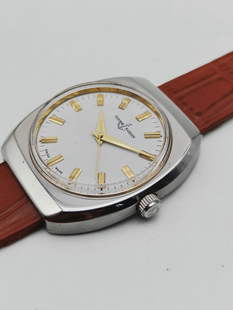 May include: A silver-toned Ulysse Nardin wristwatch with a white face and gold-colored hands and markers. The watch has a brown leather strap and a square-shaped case. The watch face displays the brand name and "Swiss Made".