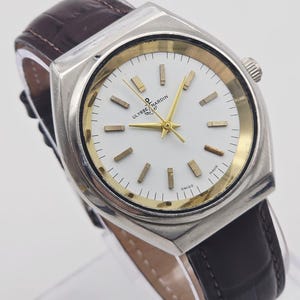 Pu&ograve; includere: Un orologio Ulysse Nardin vintage con cassa quadrata argentata e quadrante bianco. L'orologio presenta lancette e indici dorati e un cinturino in pelle marrone. Il quadrante reca la scritta "Ulysse Nardin Swiss Made".