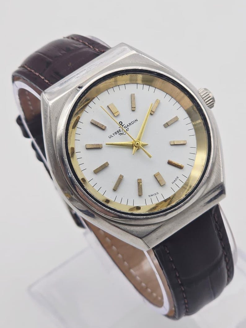 Pu&ograve; includere: Un orologio Ulysse Nardin vintage con cassa quadrata argentata e quadrante bianco. L'orologio presenta lancette e indici dorati e un cinturino in pelle marrone. Il quadrante reca la scritta "Ulysse Nardin Swiss Made".