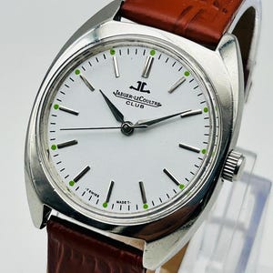 May include: A silver-toned Jaeger-LeCoultre Club watch with a white face, black hands, and green hour markers. The watch has a brown leather band. The watch face displays the Jaeger-LeCoultre logo and the words "CLUB", "T SWISS", and "MADE T."