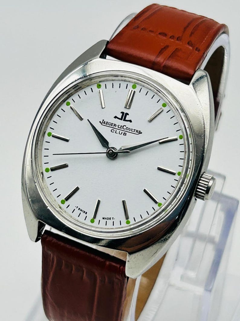 May include: A silver-toned Jaeger-LeCoultre Club watch with a white face, black hands, and green hour markers. The watch has a brown leather band. The watch face displays the Jaeger-LeCoultre logo and the words "CLUB", "T SWISS", and "MADE T."