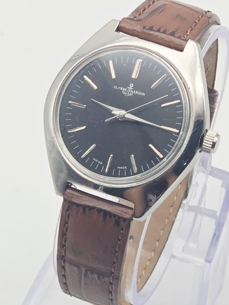 May include: A vintage Ulysse Nardin wristwatch with a dark face and silver-tone case. The watch features a brown leather band and is displayed on a clear stand. The watch face has silver markers and hands.