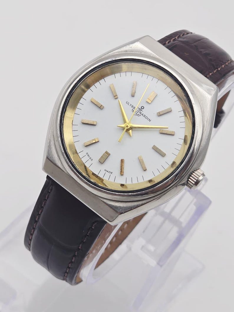 Pu&ograve; includere: Un orologio Ulysse Nardin argentato con quadrante bianco, dettagli dorati e cinturino in pelle marrone. L'orologio ha una cassa rettangolare e un design classico. Il quadrante mostra il testo "Ulysse Nardin" e "Swiss Made".