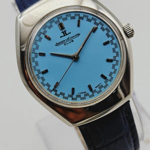 May include: A silver Jaeger-LeCoultre Club watch with a light blue face and black hands. The watch has a black leather strap and a silver case. The watch face features black rectangular markers and the Jaeger-LeCoultre logo.