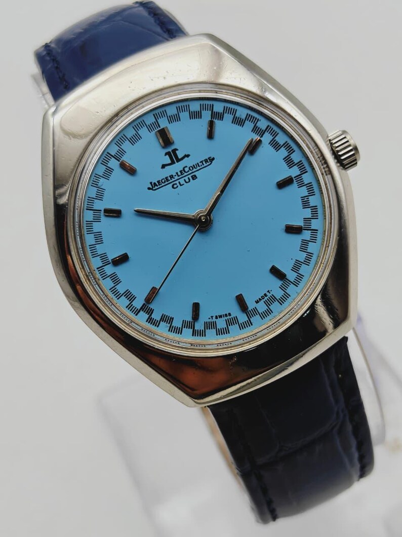 May include: A silver Jaeger-LeCoultre Club watch with a light blue face and black hands. The watch has a black leather strap and a silver case. The watch face features black rectangular markers and the Jaeger-LeCoultre logo.