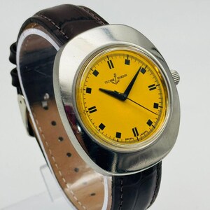May include: A vintage Ulysse Nardin wristwatch with a bright yellow face and black markers. The watch has a silver-tone case and a dark brown leather strap. The watch face displays the brand name and the words "SWISS MADE".