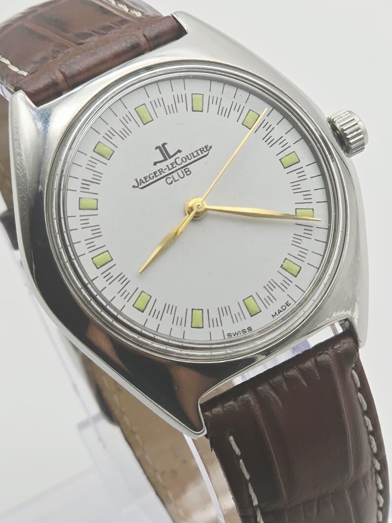 May include: A silver-toned Jaeger-LeCoultre Club watch with a white face, gold hands, and a brown leather band. The watch face features the Jaeger-LeCoultre logo and the word "CLUB". The watch has a round face and a textured brown leather band.
