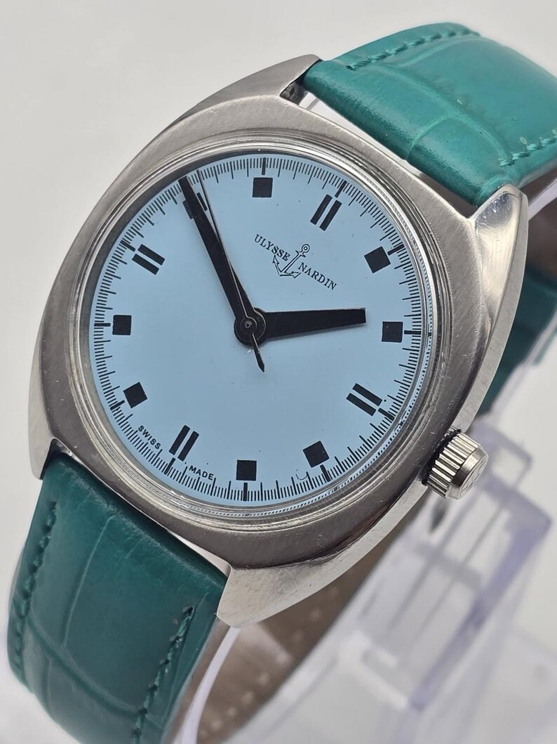 Peut inclure: Une montre-bracelet argent&eacute;e avec un cadran bleu clair et des aiguilles et index noirs. La montre a un bracelet en cuir vert. Le cadran de la montre porte les mots "Ulysse Nardin" et "Swiss Made".