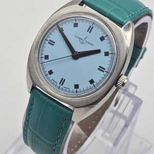 Peut inclure: Une montre Ulysse Nardin vintage avec un cadran bleu clair et des aiguilles noires. La montre a un bo&icirc;tier argent&eacute; et un bracelet en cuir vert d'eau. La montre est pr&eacute;sent&eacute;e sur un support transparent.