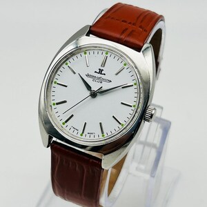 May include: A silver-toned Jaeger-LeCoultre Club watch with a white face, black hands, and a brown leather band. The watch face has green accents and the brand name is visible. The watch is displayed on a clear stand.