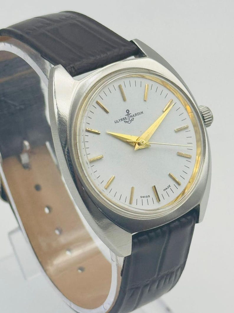 May include: A silver wristwatch with a white face and gold hands. The watch has a black leather strap and the brand name "Ulysse Nardin" on the face. The watch is a classic design.