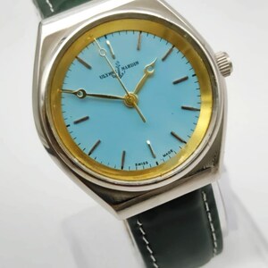 May include: A silver-toned wristwatch with a light blue face and gold-colored accents. The watch features the text "ULYSSE NARDIN SWISS MADE" and has a dark green leather band with white stitching. The watch hands are gold-colored.