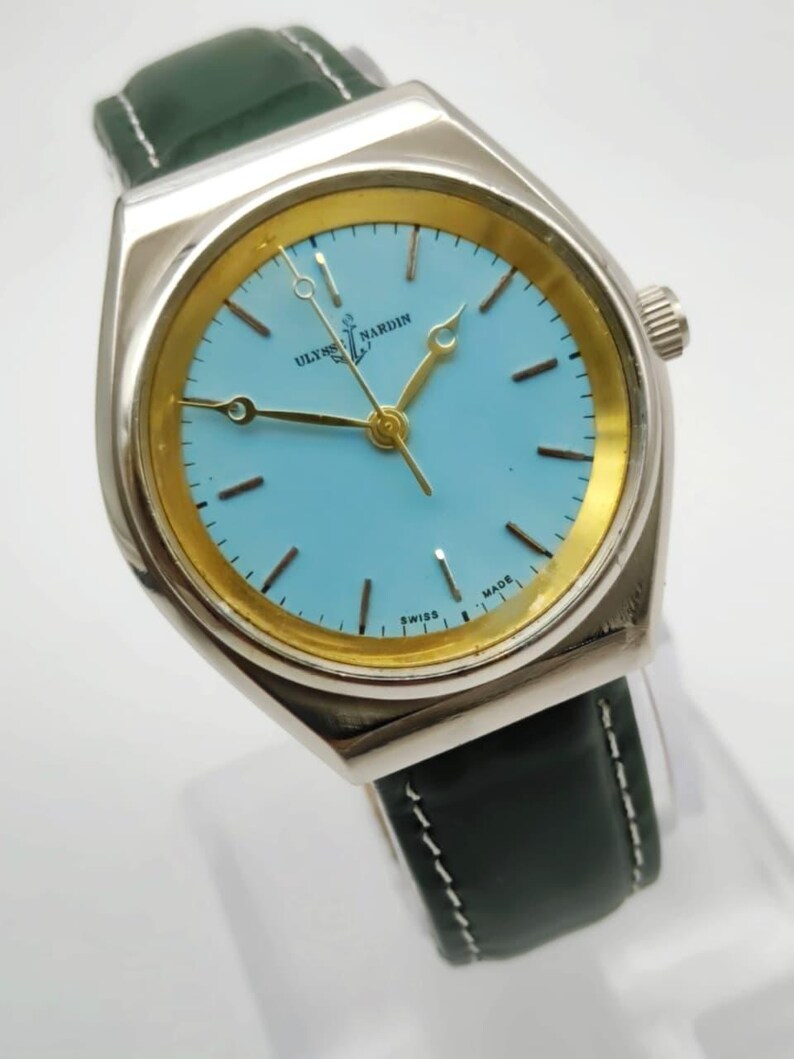May include: A silver-toned wristwatch with a light blue face and gold-colored accents. The watch features the text "ULYSSE NARDIN SWISS MADE" and has a dark green leather band with white stitching. The watch hands are gold-colored.