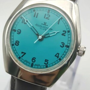 Op de afbeelding: Een zilverkleurig Jaeger-LeCoultre Club horloge met een turquoise wijzerplaat en zwarte cijfers. Het horloge heeft een bruine leren band en een zilveren lunette. De wijzerplaat toont het Jaeger-LeCoultre Club logo en de tekst "SWISS MADE".