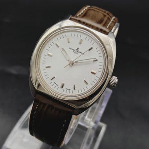 May include: A silver-tone wristwatch with a round white dial, silver hour markers, and silver hands. The watch features the Ulysse Nardin logo and anchor symbol at the top, with the text Swiss Made at the bottom. It has a brown leather strap.