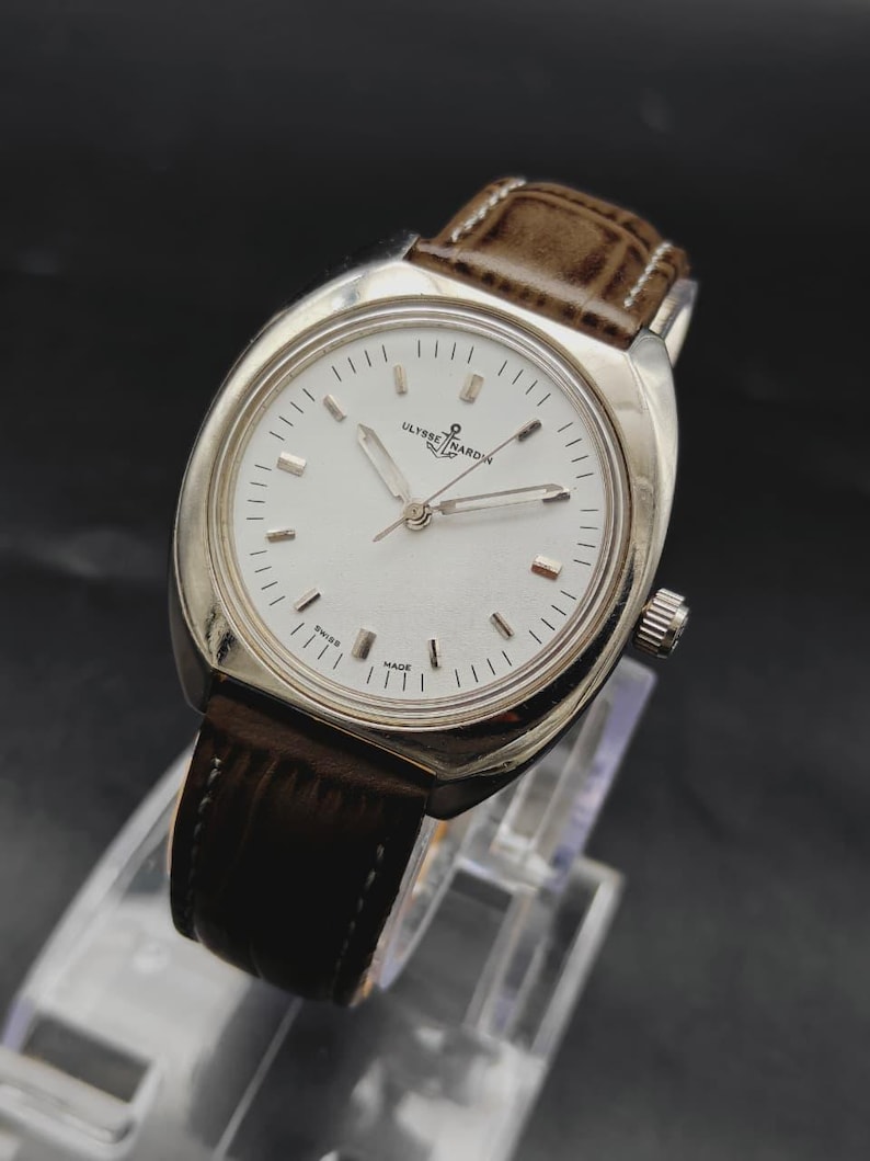 May include: A silver-tone wristwatch with a round white dial, silver hour markers, and silver hands. The watch features the Ulysse Nardin logo and anchor symbol at the top, with the text Swiss Made at the bottom. It has a brown leather strap.