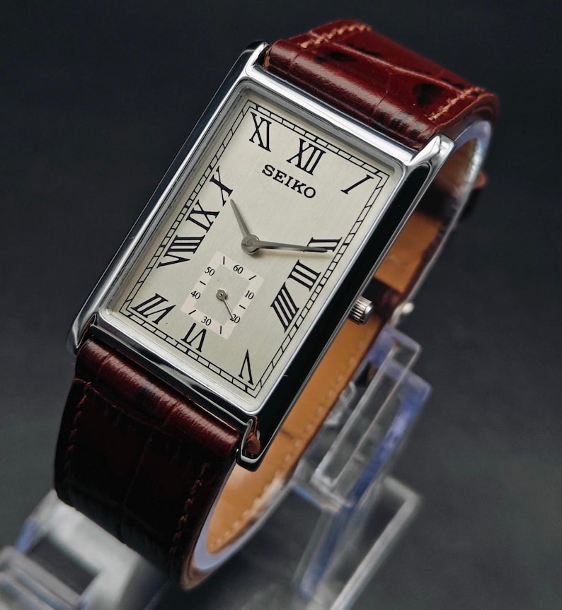 May include: A rectangular Seiko wristwatch with a silver-colored face, black Roman numerals, and a subdial. The watch has a brown leather band. The watch is displayed on a clear stand against a dark background.