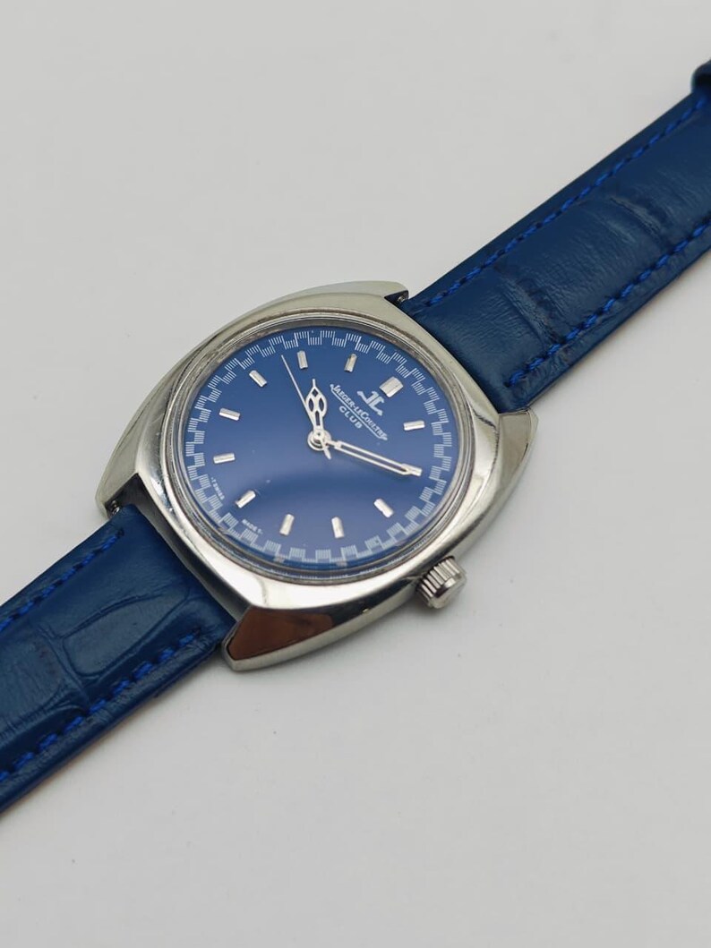 Puede incluir: Un reloj de pulsera vintage con esfera azul y caja plateada. El reloj presenta una correa de cuero azul y el texto "JAEGER-LECOULTRE CLUB" en la esfera. El reloj tiene un dise&ntilde;o cl&aacute;sico.