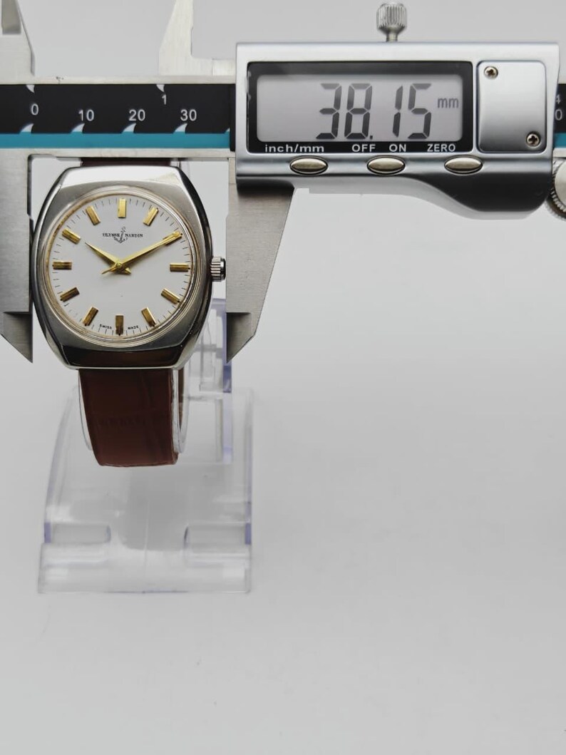 May include: A silver-toned watch with a white face and gold hands and markers. The watch has a brown leather band. The digital caliper shows the watch is 38.15 mm, or approximately 1.5 inches. The watch is labeled "OLIVER MARLEN".