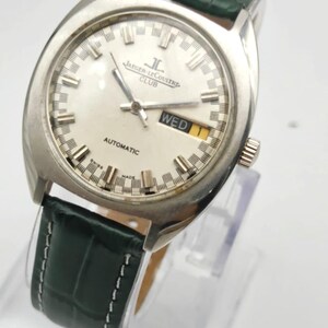 Puede incluir: Un reloj autom&aacute;tico Jaeger-LeCoultre Club vintage con esfera plateada y correa de cuero verde. La esfera muestra el d&iacute;a y la fecha, con la palabra "WED" visible. El reloj tiene una caja plateada y una correa de cuero verde texturizada.