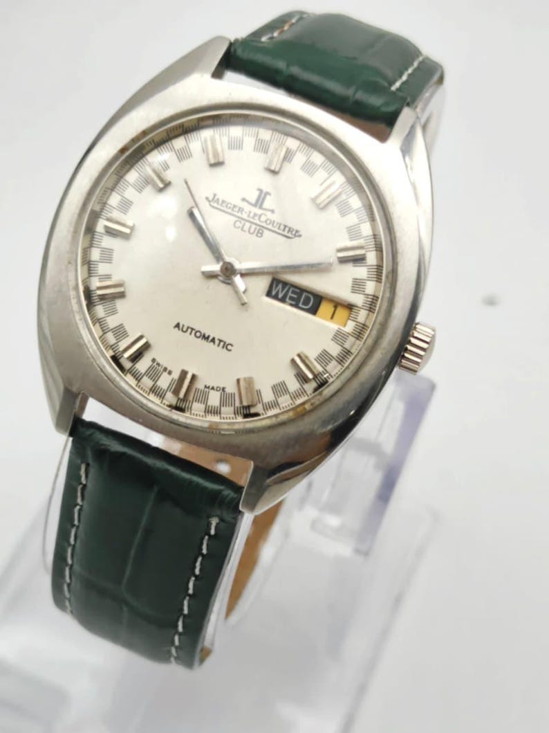 Puede incluir: Un reloj autom&aacute;tico Jaeger-LeCoultre Club vintage con esfera plateada y correa de cuero verde. La esfera muestra el d&iacute;a y la fecha, con la palabra "WED" visible. El reloj tiene una caja plateada y una correa de cuero verde texturizada.