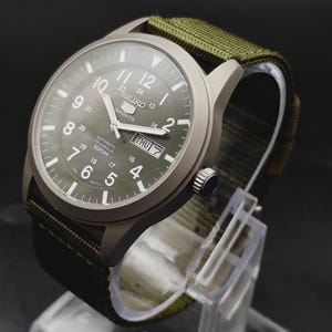 Puede incluir: Un reloj autom&aacute;tico Seiko 5 Sports con caja gris mate y correa de tela verde oliva. La esfera presenta n&uacute;meros ar&aacute;bigos blancos, un anillo interior de 24 horas, manecillas luminosas y una ventana de d&iacute;a y fecha en la posici&oacute;n de las 3.