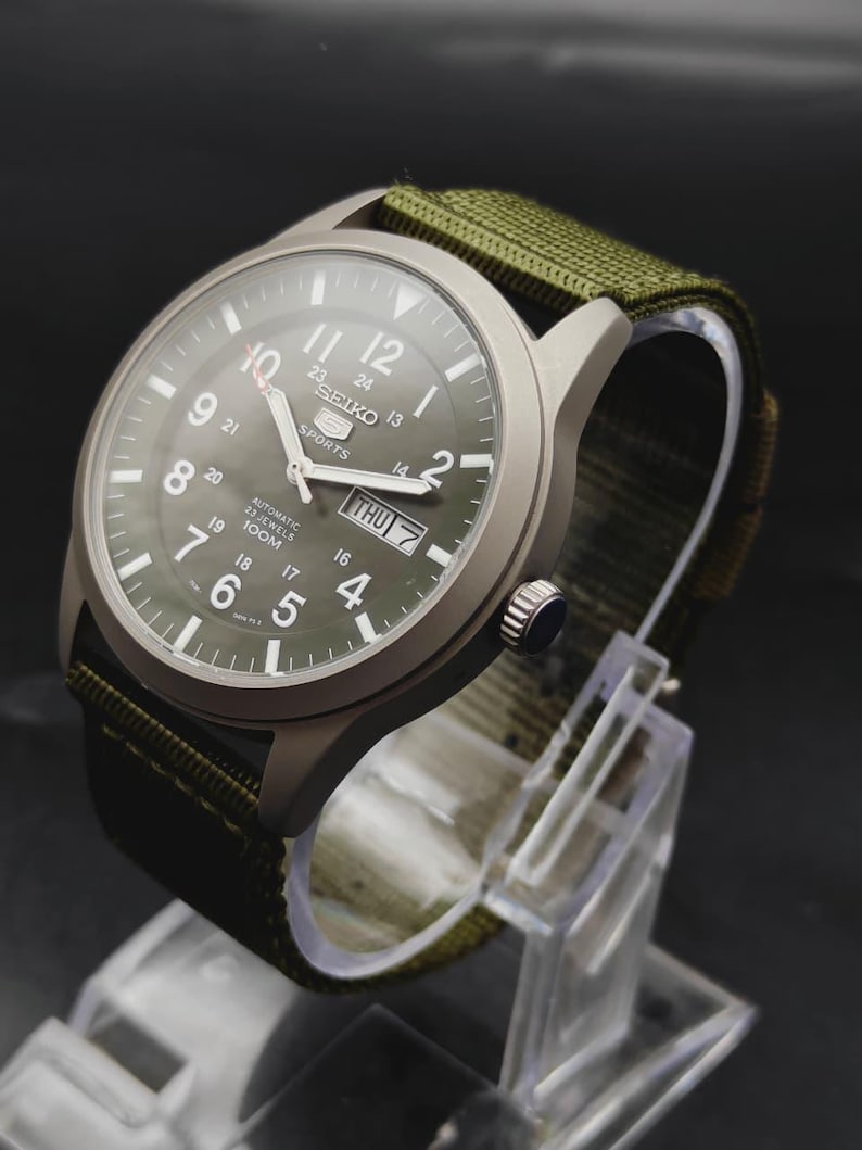 Puede incluir: Un reloj autom&aacute;tico Seiko 5 Sports con caja gris mate y correa de tela verde oliva. La esfera presenta n&uacute;meros ar&aacute;bigos blancos, un anillo interior de 24 horas, manecillas luminosas y una ventana de d&iacute;a y fecha en la posici&oacute;n de las 3.