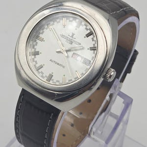 Puede incluir: Un reloj autom&aacute;tico Jaeger-LeCoultre Club plateado con esfera blanca, visualizaci&oacute;n de d&iacute;a y fecha, y correa de cuero negro. El reloj tiene una caja rectangular y una correa de cuero negro texturizado. El reloj se exhibe sobre un soporte transparente.