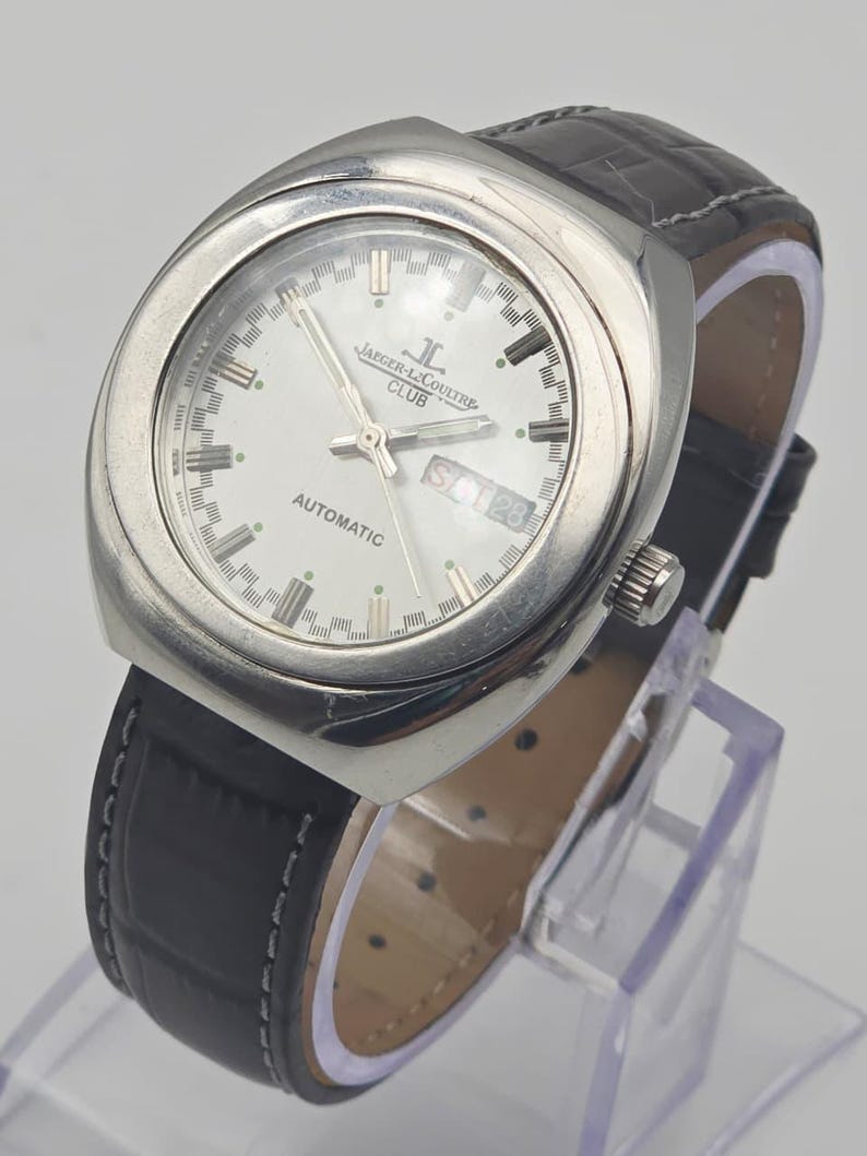 Puede incluir: Un reloj autom&aacute;tico Jaeger-LeCoultre Club plateado con esfera blanca, visualizaci&oacute;n de d&iacute;a y fecha, y correa de cuero negro. El reloj tiene una caja rectangular y una correa de cuero negro texturizado. El reloj se exhibe sobre un soporte transparente.