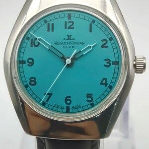 Op de afbeelding: Een zilverkleurig Jaeger-LeCoultre Club horloge met een turquoise wijzerplaat en een zwarte leren band. De wijzerplaat heeft witte cijfers en wijzers, met de merknaam en de tekst "SWISS MADE". Het horloge heeft een ronde wijzerplaat en een rechthoekige kast.