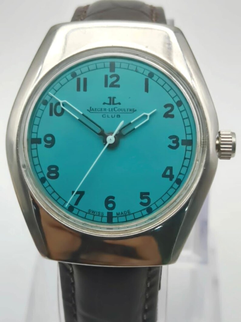 Op de afbeelding: Een zilverkleurig Jaeger-LeCoultre Club horloge met een turquoise wijzerplaat en een zwarte leren band. De wijzerplaat heeft witte cijfers en wijzers, met de merknaam en de tekst "SWISS MADE". Het horloge heeft een ronde wijzerplaat en een rechthoekige kast.