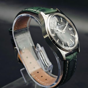 K&ouml;nnte beinhalten: Eine silberfarbene Armbanduhr mit einem dunkelgrauen Zifferblatt und einem gr&uuml;nen Lederarmband. Das Zifferblatt der Uhr hat silberne Stundenmarkierungen und Zeiger. Das Armband ist aus strukturiertem gr&uuml;nem Leder mit wei&szlig;en N&auml;hten. Die Uhr wird auf einem transparenten St&auml;nder pr&auml;sentiert.