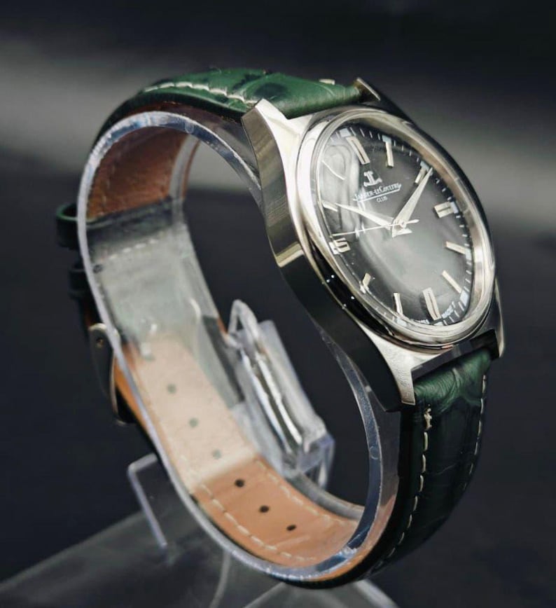 K&ouml;nnte beinhalten: Eine silberfarbene Armbanduhr mit einem dunkelgrauen Zifferblatt und einem gr&uuml;nen Lederarmband. Das Zifferblatt der Uhr hat silberne Stundenmarkierungen und Zeiger. Das Armband ist aus strukturiertem gr&uuml;nem Leder mit wei&szlig;en N&auml;hten. Die Uhr wird auf einem transparenten St&auml;nder pr&auml;sentiert.