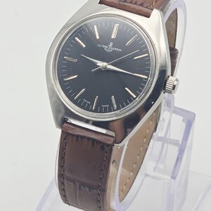 May include: A vintage Ulysse Nardin wristwatch with a black dial and silver hands and markers. The watch has a stainless steel case and a brown leather strap. The watch is displayed on a clear stand.
