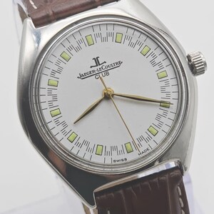 May include: A silver Jaeger-LeCoultre Club watch with a white face, gold hands, and a brown leather strap. The watch face features the Jaeger-LeCoultre logo and the word "CLUB". The watch is marked "SWISS MADE".