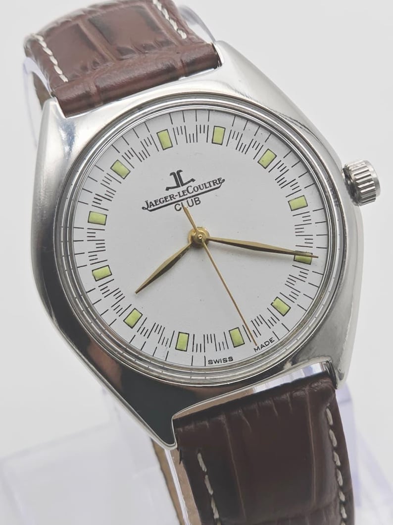 May include: A silver Jaeger-LeCoultre Club watch with a white face, gold hands, and a brown leather strap. The watch face features the Jaeger-LeCoultre logo and the word "CLUB". The watch is marked "SWISS MADE".