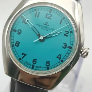 Op de afbeelding: Een vintage Jaeger-LeCoultre Club horloge met een turquoise wijzerplaat en een zilverkleurige kast. Het horloge heeft zwarte cijfers, witte wijzers en een bruine leren band. De tekst "JAEGER-LECOULTRE CLUB" is zichtbaar. Swiss Made.