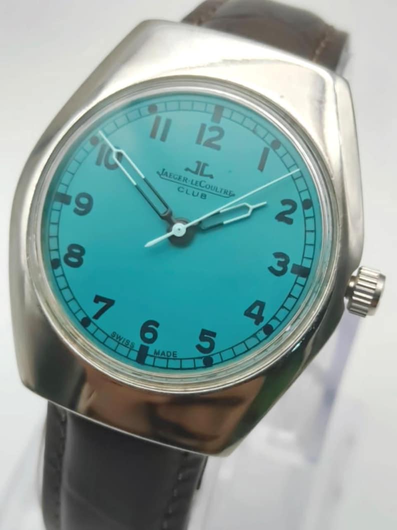 Op de afbeelding: Een vintage Jaeger-LeCoultre Club horloge met een turquoise wijzerplaat en een zilverkleurige kast. Het horloge heeft zwarte cijfers, witte wijzers en een bruine leren band. De tekst "JAEGER-LECOULTRE CLUB" is zichtbaar. Swiss Made.