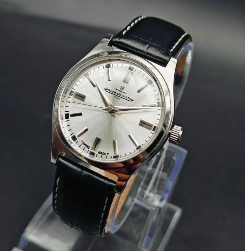 May include: A silver-toned wristwatch with a black leather band. The watch face is silver with black markers and hands. The watch is displayed on a clear stand against a dark background. The watch has the text "Jaeger-LeCoultre Club" on the face.