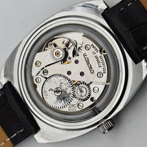 May include: Close-up of a vintage silver wristwatch with a black leather strap. The watch face is transparent, revealing the intricate mechanical movement with gears, jewels, and the text "SWISS SEVENTEEN 17 JEWELS LE COULTRE CO".