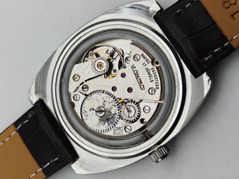 May include: Close-up of a vintage silver wristwatch with a black leather strap. The watch face is transparent, revealing the intricate mechanical movement with gears, jewels, and the text "SWISS SEVENTEEN 17 JEWELS LE COULTRE CO".