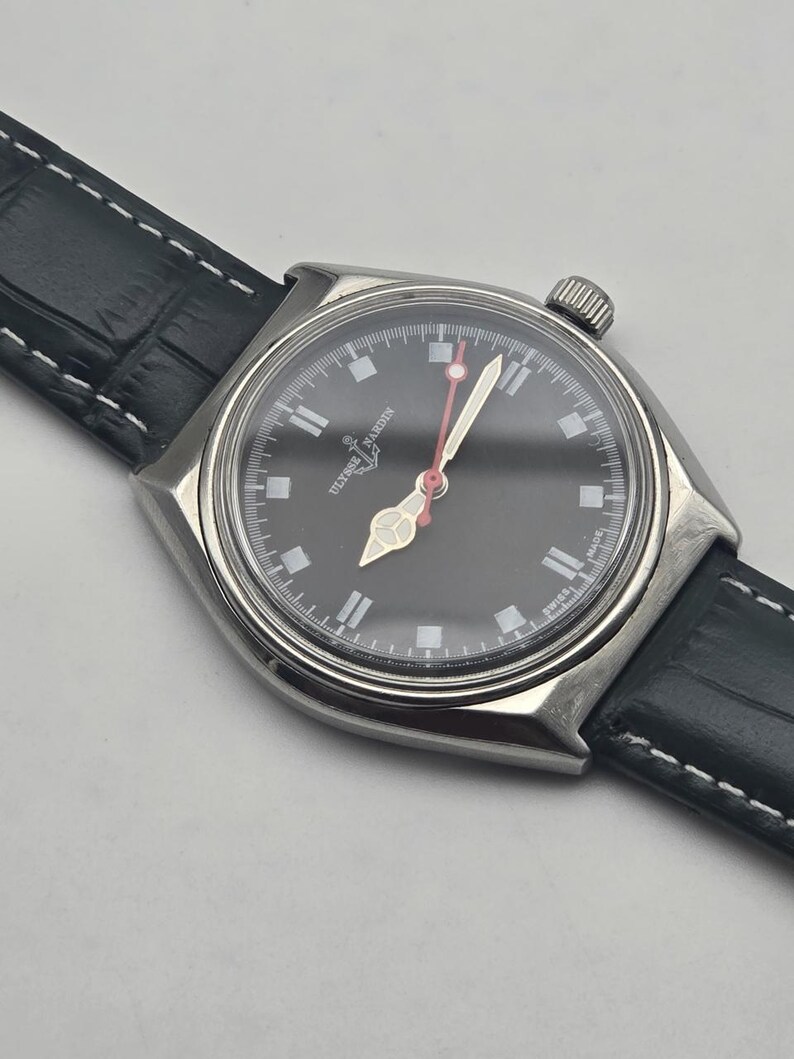 Puede incluir: Un reloj de pulsera de acero inoxidable con esfera negra y correa de cuero negro. La esfera del reloj presenta marcadores de hora y agujas blancas, con un segundero rojo. El reloj est&aacute; marcado "SWISS MADE".