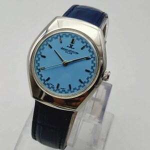 May include: A silver Jaeger-LeCoultre Club watch with a light blue face and a dark blue leather strap. The watch face has a checkered pattern around the edge and the text "JAEGER-LECOULTRE CLUB".