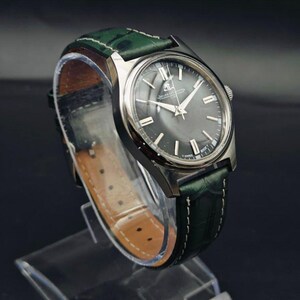 K&ouml;nnte beinhalten: Eine silberfarbene Armbanduhr mit dunkelgrauem Zifferblatt und silbernen Zeigern. Das gr&uuml;ne Lederarmband hat wei&szlig;e N&auml;hte und eine braune Unterseite. Die Uhr wird auf einem transparenten St&auml;nder pr&auml;sentiert.