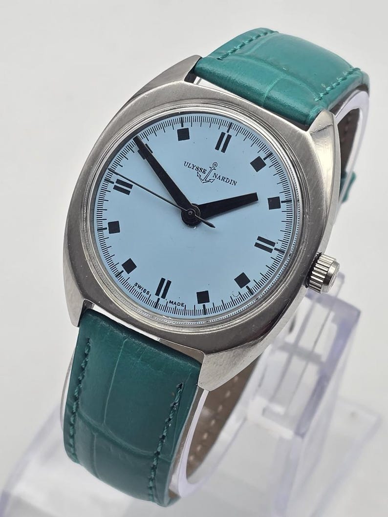 Peut inclure: Une montre argent&eacute;e avec un cadran bleu clair et des aiguilles noires. La montre porte l'inscription "ULYSSE NARDIN" et un bracelet en cuir vert. Le cadran comporte des index noirs rectangulaires.