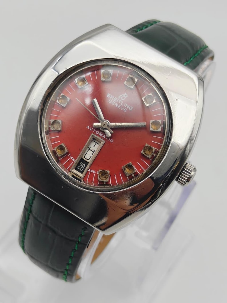 May include: A vintage Breitling Geneve automatic watch with a red dial and silver-tone case. The watch features a black leather strap with green stitching. The dial includes the date and hour markers. The watch is displayed on a clear stand.