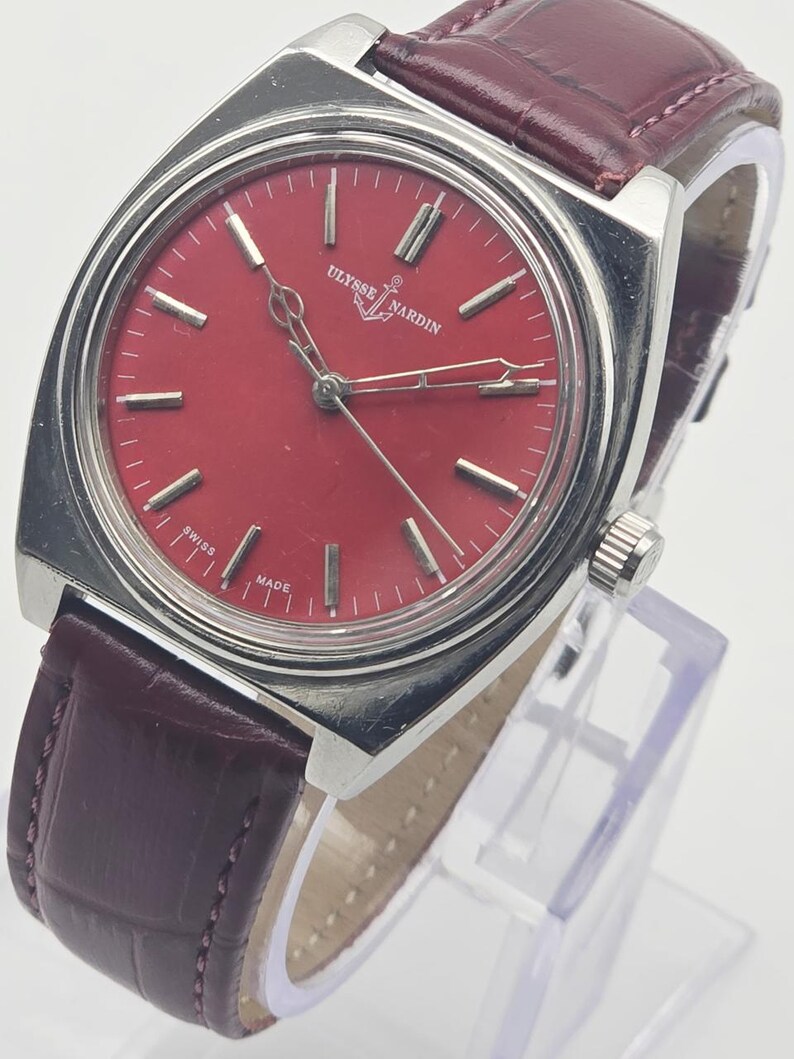 May include: A vintage Ulysse Nardin wristwatch with a red dial and silver-tone case. The watch features a burgundy leather strap. The dial has silver markers and hands. The watch is marked "Swiss Made".