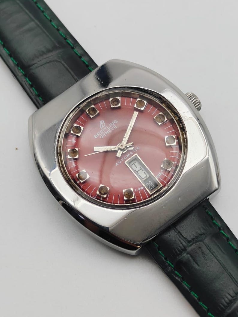 May include: A vintage Breitling Gen&egrave;ve automatic watch with a red dial and a date display. The watch has a silver-toned square case and a black leather strap with green stitching. The watch face features silver hands and hour markers.